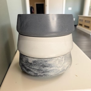 Op de afbeelding: Drie gestapelde, ronde betonnen kommen in grijstinten, wit en marmer. De bovenste kom is effen donkergrijs, de middelste is wit en de onderste kom heeft een gemarmerd grijs-wit patroon. De kommen zijn gestapeld op een wit oppervlak.