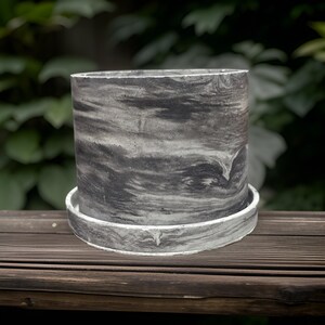 May include: A grey concrete planter with a marbled pattern. The planter has a wide, flat base and a cylindrical shape.