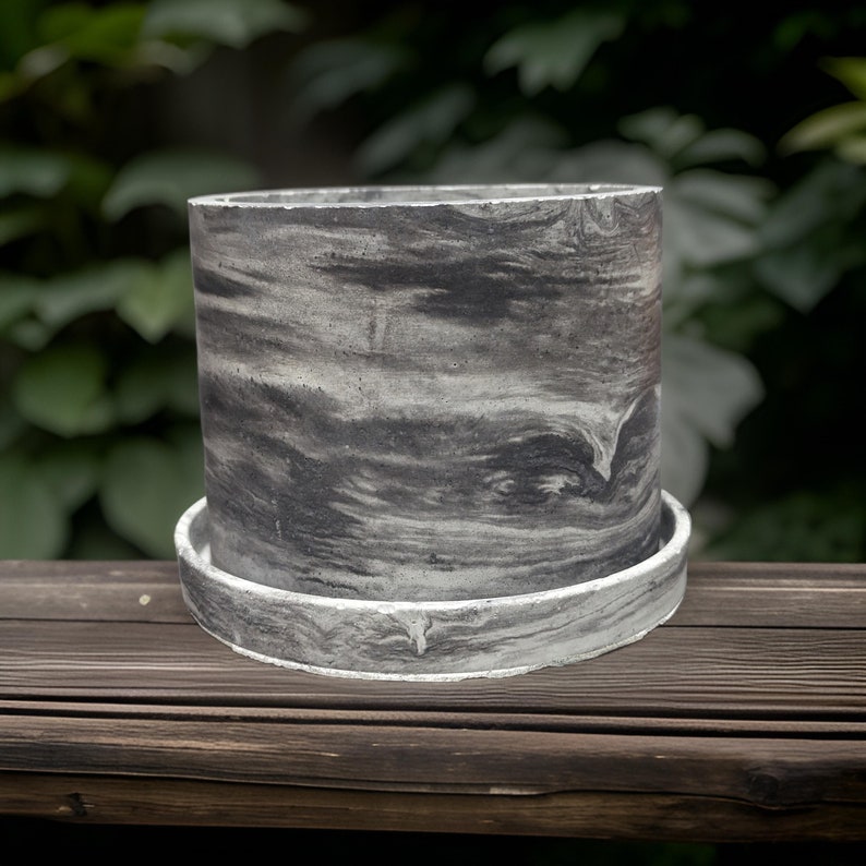 May include: A grey concrete planter with a marbled pattern. The planter has a wide, flat base and a cylindrical shape.