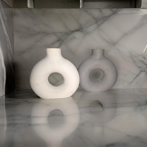 May include: Two ceramic vases with a donut-shaped opening. The vase on the left is white and the vase on the right is grey. The vases are sitting on a marble surface.