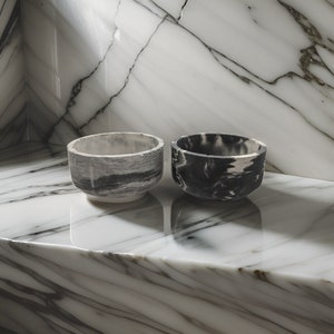 Puede incluir: Dos cuencos pequeños hechos de hormigón con un patrón de mármol. Un cuenco es blanco y negro, y el otro es gris y blanco. Los cuencos están sobre una superficie de mármol blanco.