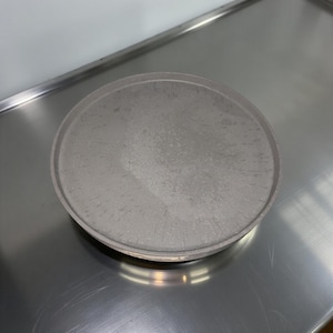May include: A round, gray metal tray with a slightly textured surface. The tray has a raised edge and sits on a reflective, metallic surface. The tray is circular and appears to be made of a solid material.