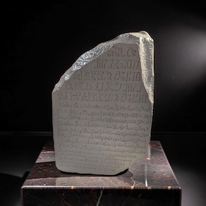 May include: A translucent, light-colored stone slab with a jagged, textured top edge. The flat front surface is engraved with multiple rows of ancient-style script characters. The stone has a semi-transparent quality and a smooth, polished face.