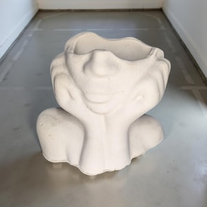 May include: White ceramic planter shaped like a human head with hands resting on the chin. The planter has a smooth, textured surface and a wide opening at the top.