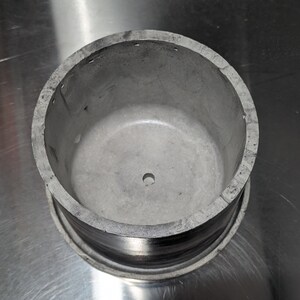 May include: A silver metal container with a round opening and a small hole in the centre. The container has a smooth, slightly textured surface.