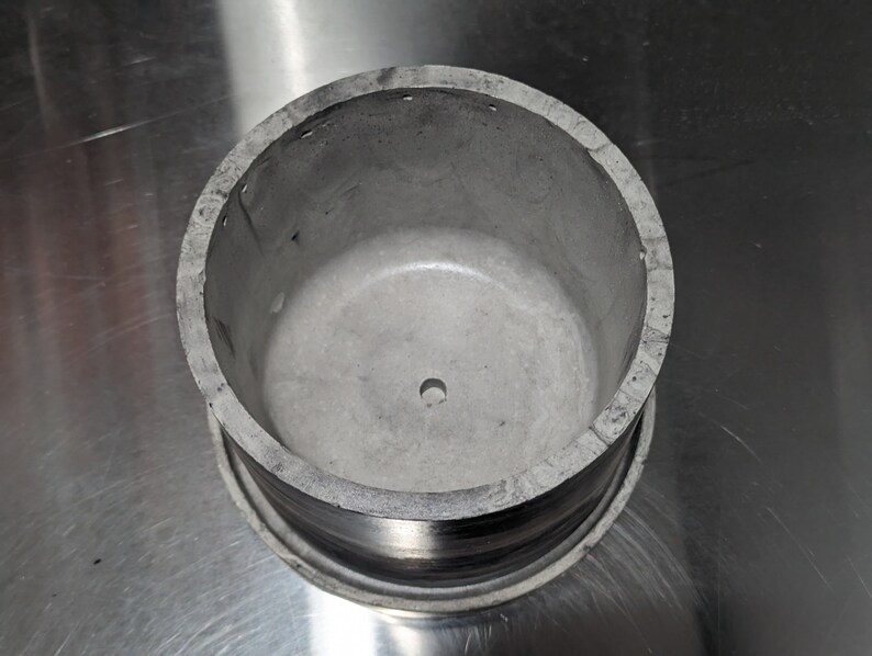May include: A silver metal container with a round opening and a small hole in the centre. The container has a smooth, slightly textured surface.