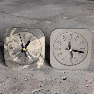 Puede incluir: Dos relojes cuadrados de hormigón con manecillas negras y detalles dorados. Uno tiene una esfera gris y blanca veteada, el otro es gris liso. Los relojes tienen un diseño minimalista y están sobre un fondo gris.