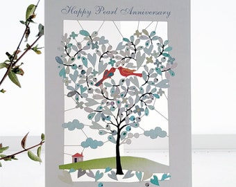 May include: A white card with the text "Happy Pearl Anniversary" at the top. The card features a heart-shaped tree with two red birds perched on a branch. The tree is surrounded by blue and white clouds and a small green hill with a red house.