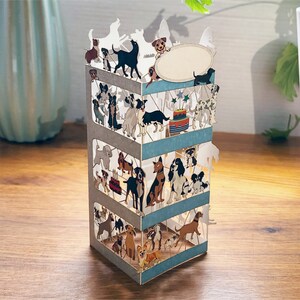 Puede incluir: Una tarjeta pop-up 3D con un dise&ntilde;o de varios niveles que presenta un pastel de cumplea&ntilde;os y muchos perros. La tarjeta est&aacute; hecha de papel y tiene una combinaci&oacute;n de colores azul claro, gris y blanco.