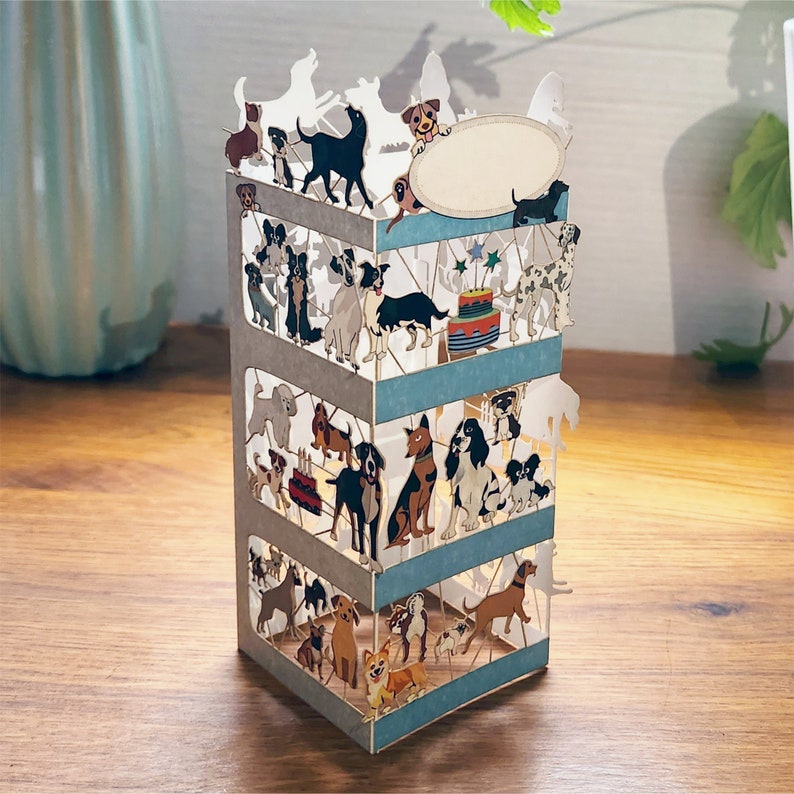 Puede incluir: Una tarjeta pop-up 3D con un dise&ntilde;o de varios niveles que presenta un pastel de cumplea&ntilde;os y muchos perros. La tarjeta est&aacute; hecha de papel y tiene una combinaci&oacute;n de colores azul claro, gris y blanco.