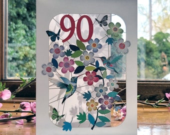 May include: A white card with a cut-out design featuring a number "90" in pink, two hummingbirds, butterflies, and colorful flowers. The card is on a wooden surface.