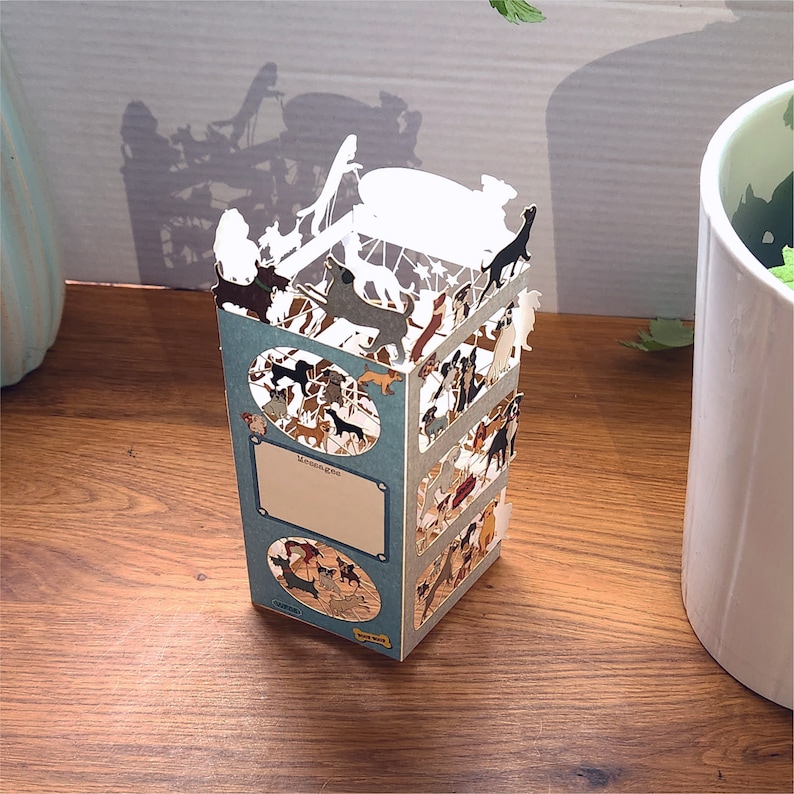 Puede incluir: Una tarjeta pop-up 3D con una variedad de razas de perros en diferentes poses. La tarjeta est&aacute; hecha de papel y tiene un fondo azul con un borde blanco. La tarjeta est&aacute; doblada y de pie sobre una superficie de madera. La tarjeta tiene las palabras "El amor es una palabra de cuatro patas" impresas en ella.
