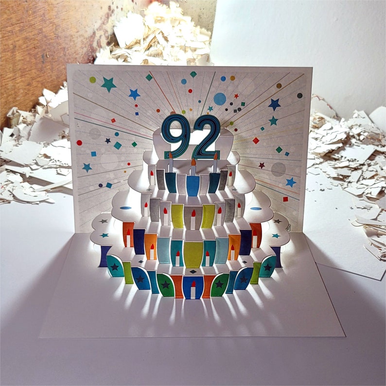 May include: A pop-up card with a birthday cake design. The cake is decorated with colorful candles and the number "92" on top. The card is white with a colorful background.