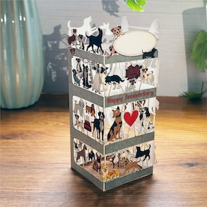 May include: A 3D pop-up card with a grey background and a white frame. The card features a variety of dogs in different poses and colours. The card says "Happy Anniversary" in red letters.