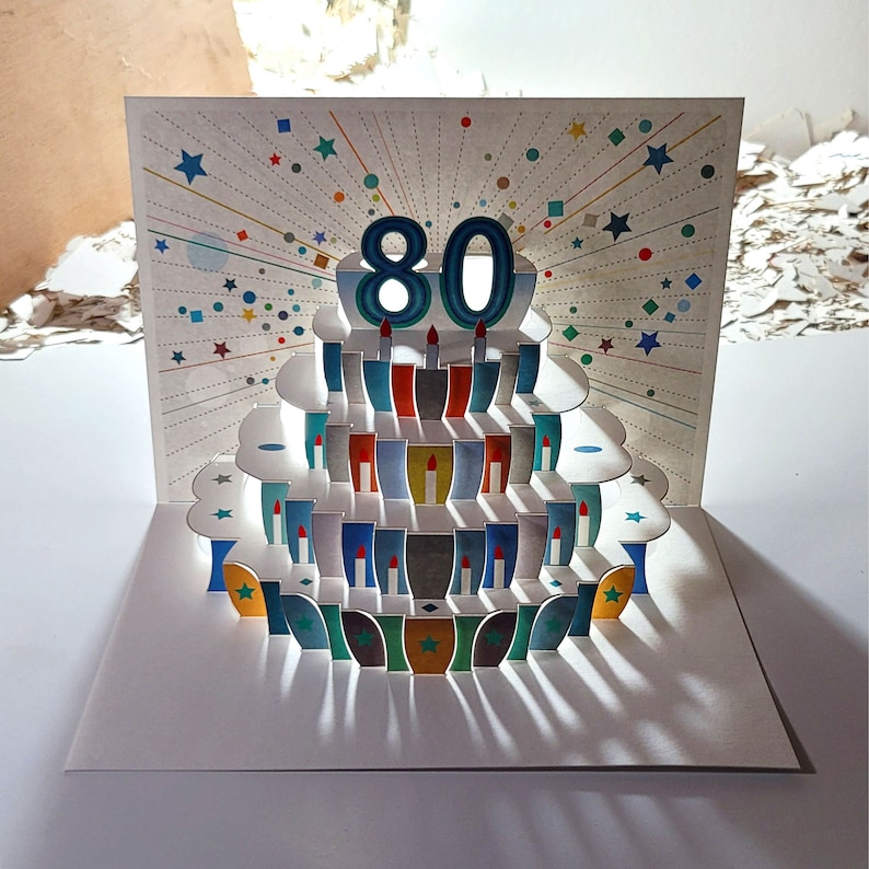 May include: A pop-up card featuring a colourful cake design. The cake has 80 candles and is decorated with blue, green and red accents. The card is ideal for celebrating a birthday.