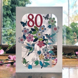 May include: A white card with a cut-out design featuring a number 80, colorful flowers, and hummingbirds. The card is perfect for an 80th birthday celebration.