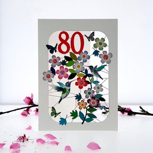 May include: A white card with a red number 80 in a window with a floral design and hummingbirds. The flowers are pink, purple, blue, and green.
