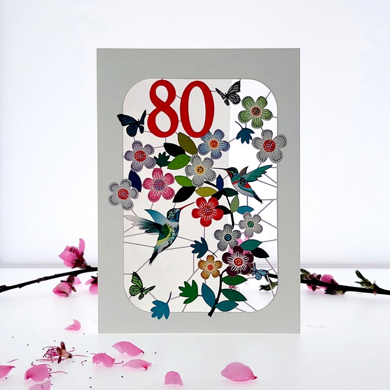 May include: A white card with a red number 80 in a window with a floral design and hummingbirds. The flowers are pink, purple, blue, and green.