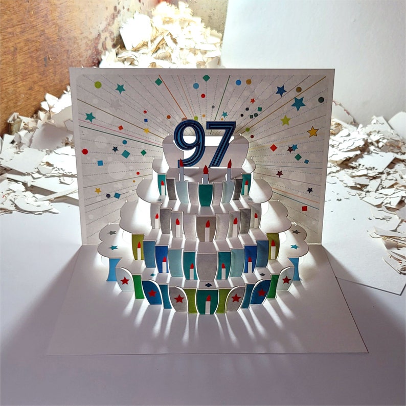 May include: A 3D pop-up card with a birthday cake design. The cake is decorated with blue and white frosting and has 97 candles on top. The card is white with a colorful confetti design.