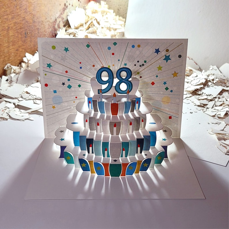 May include: A white pop-up card with a colorful layered cake design. The cake has blue and white frosting and the number 98 on top. The card is decorated with colorful confetti and stars.