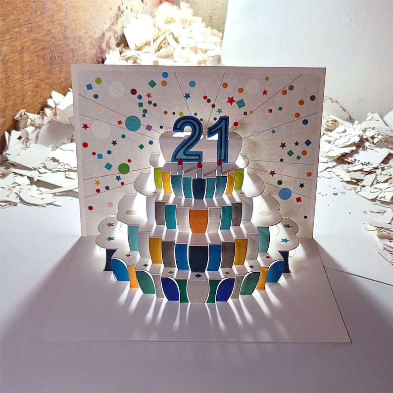 May include: A pop-up card with a colorful cake design. The cake has a blue and white number 21 on top. The card is decorated with confetti and stars.