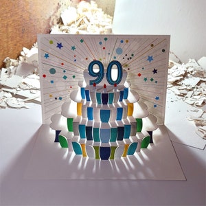 May include: A pop-up card with a colorful cake design. The cake has a blue and white striped base and is decorated with a blue number "90" on top. The card is perfect for celebrating a 90th birthday.