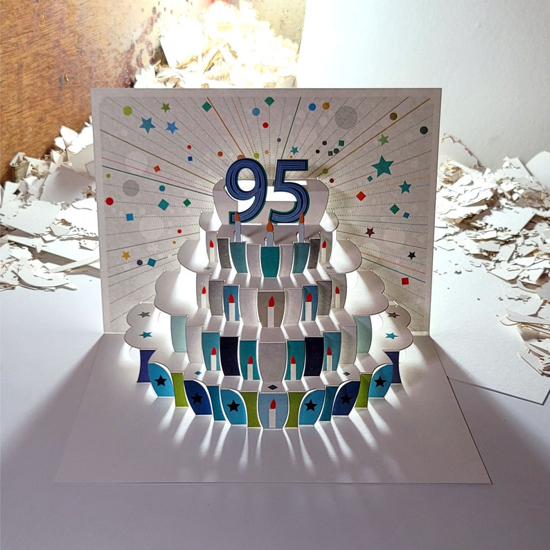 May include: A 3D pop-up card featuring a birthday cake with 95 candles. The cake is blue, green, and white with a white background. The card is perfect for celebrating a 95th birthday.