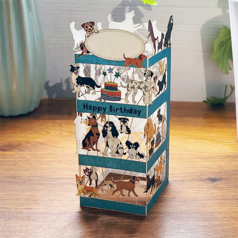 Puede incluir: Una tarjeta pop-up 3D con fondo azul y una variedad de perros en diferentes poses. La tarjeta dice "Feliz cumplea&ntilde;os" y presenta un pastel con velas.