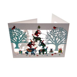 May include: A pop-up card featuring a Christmas tree made of elephants wearing party hats. The elephants are holding onto each other's tails and are decorated with colourful patterns. The card is set against a white background with blue trees on either side.