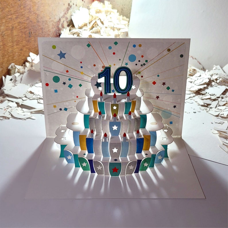 May include: A pop-up card with a colorful cake design. The cake has a blue number 10 on top and is surrounded by colorful candles. The card is decorated with confetti and stars.