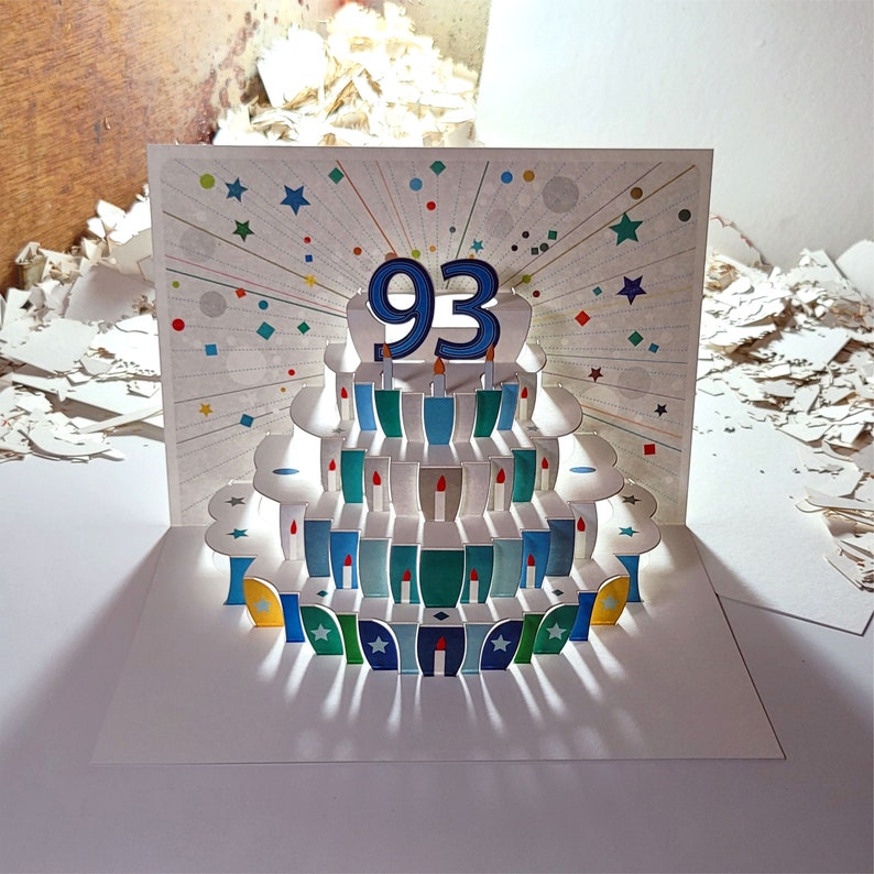 May include: A white pop-up card with a blue and white cake with candles and the number 93 on top. The cake is surrounded by a colorful confetti design.