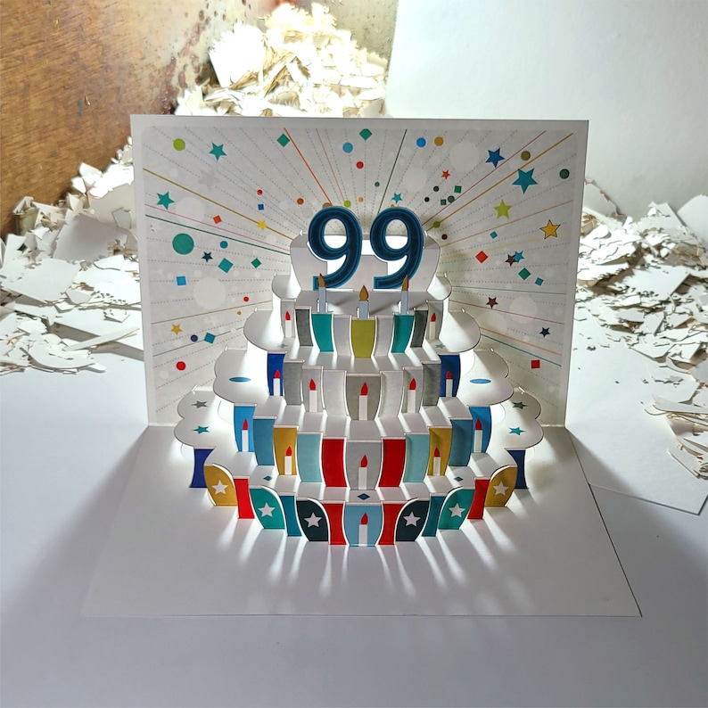 May include: A pop-up card with a colorful cake design. The cake has 99 candles and is decorated with red, blue, and green frosting. The card is perfect for a 99th birthday celebration.