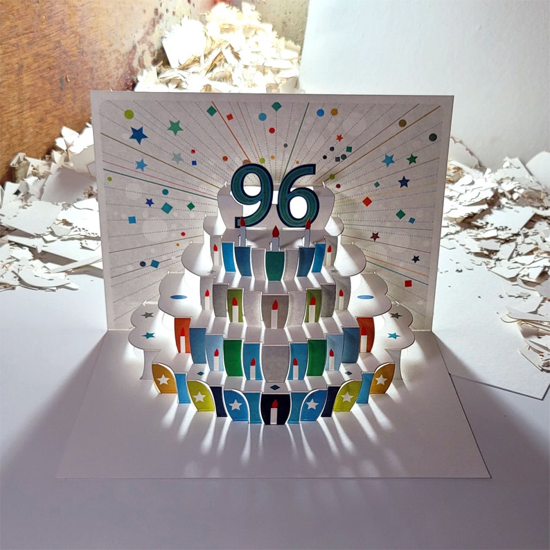 May include: A white pop-up card with a 3D cake design. The cake has 96 candles and is decorated with colorful details. The card is perfect for a 96th birthday celebration.