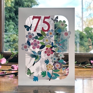 May include: A white card with a cut-out design featuring a hummingbird, butterflies, and flowers. The number 75 is displayed in red on the card.