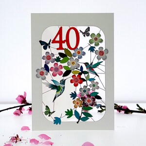 Puede incluir: Una tarjeta blanca con el n&uacute;mero "40" en rojo en una ventana recortada. La ventana muestra un dise&ntilde;o colorido de flores, hojas y colibr&iacute;es.