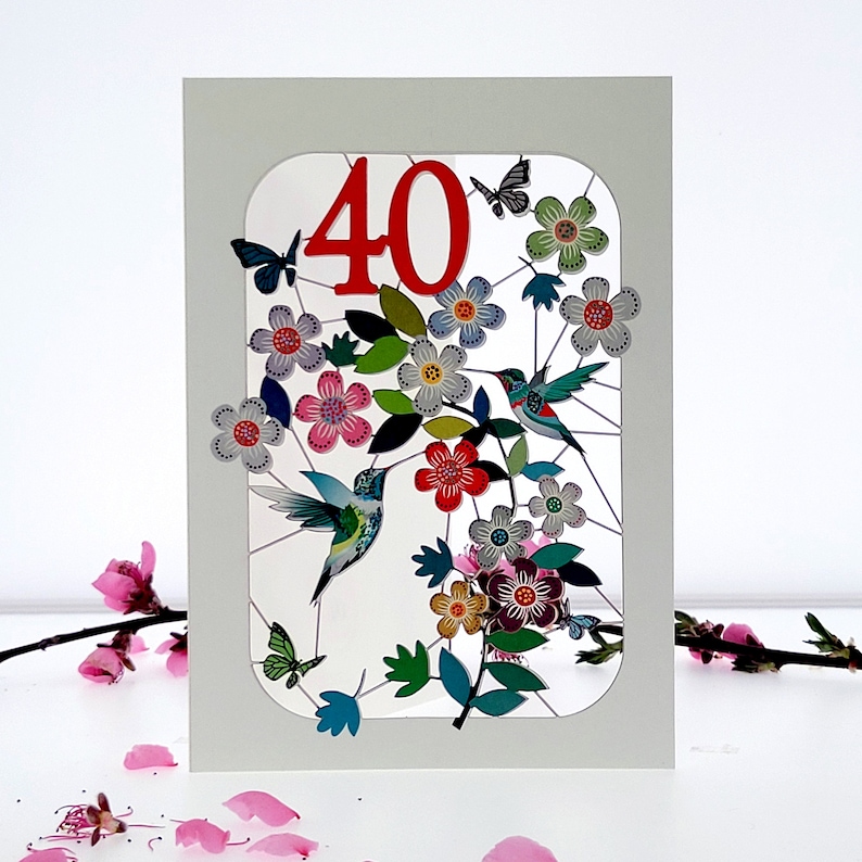 Puede incluir: Una tarjeta blanca con el n&uacute;mero "40" en rojo en una ventana recortada. La ventana muestra un dise&ntilde;o colorido de flores, hojas y colibr&iacute;es.