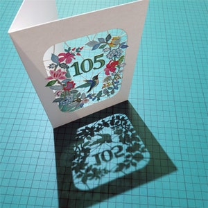 May include: A white card with a cut-out design of flowers, leaves, and a hummingbird. The number 105 is visible in the design. The card is casting a shadow on a blue and white checkered surface.