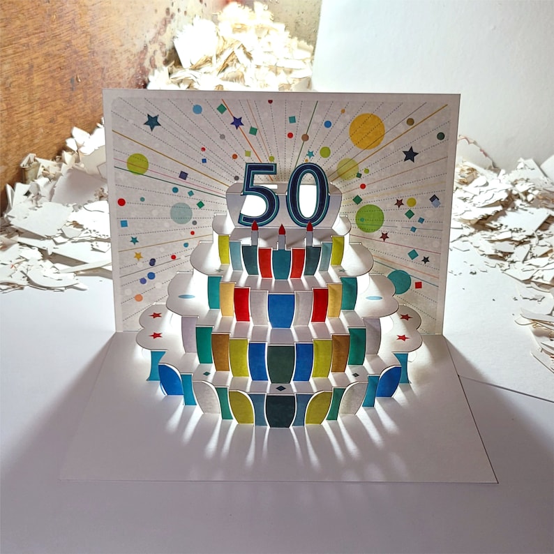 May include: A pop-up card with a colorful cake design. The cake has a number "50" on top and is decorated with colorful layers. The card is perfect for a 50th birthday celebration.