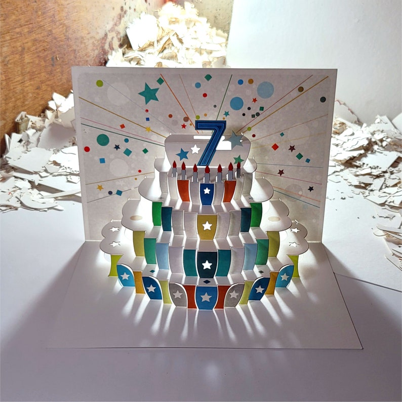 May include: A pop-up card with a colorful, layered cake design. The cake has a blue number seven on top and white stars on each layer. The card is decorated with confetti and stars.
