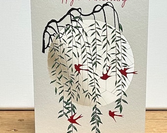 May include: A white card with the text "Happy Anniversary" in red lettering. The card features a black silhouette of a weeping willow tree with red birds flying around a white circle.