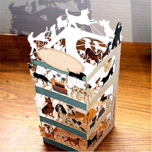 Puede incluir: Una tarjeta pop-up 3D con un carrusel blanco con una variedad de perros en diferentes poses. Los perros son todos en tonos de marr&oacute;n, negro y blanco. La tarjeta est&aacute; hecha de papel y tiene una base a rayas azul y blanca.