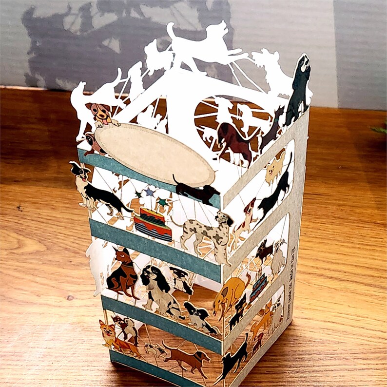 Puede incluir: Una tarjeta pop-up 3D con un carrusel blanco con una variedad de perros en diferentes poses. Los perros son todos en tonos de marr&oacute;n, negro y blanco. La tarjeta est&aacute; hecha de papel y tiene una base a rayas azul y blanca.