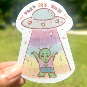 May include: A sticker featuring a cartoon alien being beamed up by a UFO. The UFO has the words "TAKE ME NOW" on it. The alien is green and wearing a blue shirt and shorts. The background is a gradient of pastel colors.