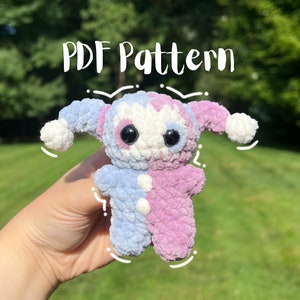 May include: A crocheted plush toy with a blue and pink color scheme. The toy has large eyes and a jester hat. The toy is being held in a hand. The text "PDF Pattern" is visible above the toy.