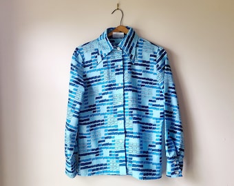 May include: A long-sleeved button-down shirt with a geometric pattern in shades of blue. The shirt is hanging on a wooden hanger against a plain white wall. The pattern consists of rectangular shapes in varying shades of blue.