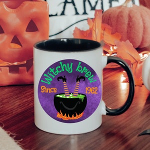 May include: White ceramic mug with a black handle. The mug has a purple and green graphic of a witch's legs sticking out of a cauldron with the text "Witchy Brew! Since 1962".