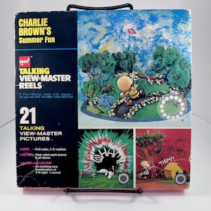 May include: A vintage Charlie Brown's Summer Fun View-Master reel set. The box features colorful illustrations of Charlie Brown and Snoopy. The set includes 21 talking View-Master pictures. The box is displayed on a black metal stand.