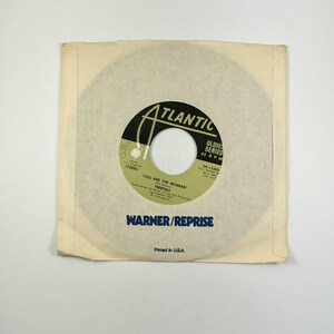 May include: A vintage 45 RPM vinyl record in a paper sleeve. The record label is black and green with the text "ATLANTIC" and "YOU ARE THE WOMAN" by Firefall. The sleeve is off-white with "WARNER/REPRISE" printed on it.