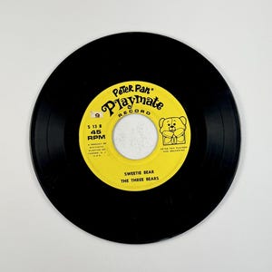 May include: A black vinyl record with a yellow label featuring the text "Peter Pan Playmate Record". The label also includes the text "Sweetie Bear The Three Bears" and a cartoon bear illustration. The record is a 45 RPM.