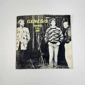 May include: Album cover for Genesis' "Throwing It All Away." The cover features three men in front of a dark wooden backdrop. The band's name is in yellow block letters above the song title.